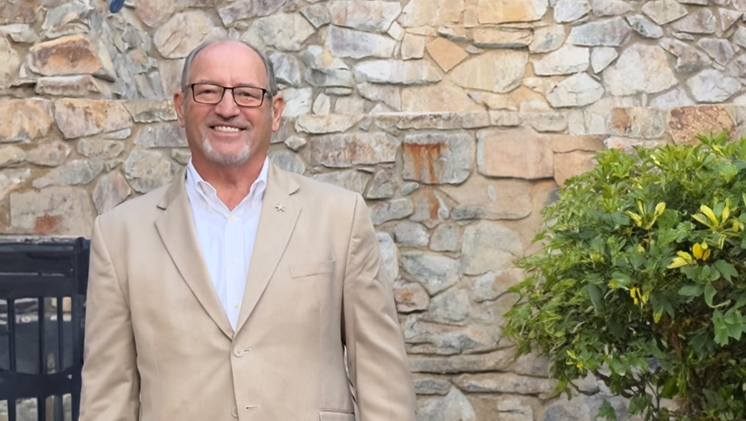 Man in beige suit smiling by stone wall and plant.
